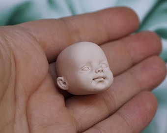 Könnte beinhalten: Ein kleiner, blasser Puppenkopf mit mattem Finish und modellierten Gesichtsdetails, darunter geschlossene Augen, eine kleine Nase und ein neutraler Mundausdruck. Der Kopf ist glatt, kahl und abgerundet, mit deutlich erkennbaren Ohren.