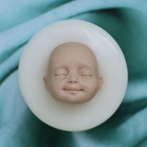 Könnte beinhalten: Eine weiße Silikonform zum Herstellen eines Baby-Puppenkopfes. Die Form ist wie ein Babygesicht geformt mit geschlossenen Augen und einem lächelnden Mund.