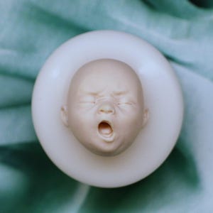 Könnte beinhalten: Eine detaillierte, blasse Skulptur eines Babykopfes mit geschlossenen Augen und offenem Mund, eingebettet in einer weißen, kreisförmigen Form. Das Gesicht des Babys zeigt einen Ausdruck von Not. Der Hintergrund ist ein weicher, blaugrüner Stoff.