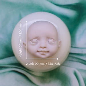 Könnte beinhalten: Ein blasses, unbemaltes Puppengesicht, 34 mm hoch und 29 mm breit, ist auf einem weißen, abgerundeten Sockel zentriert. Das Gesicht hat geschlossene Augen und ein leichtes Lächeln. Der Hintergrund ist ein weicher, türkisfarbener Stoff.