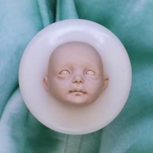 May include: A detailed, pale-toned baby doll head, set in a white, circular mould. The doll head has closed eyes, a small nose, and a defined mouth. The background is a soft, light green fabric, providing a gentle contrast to the doll's features.