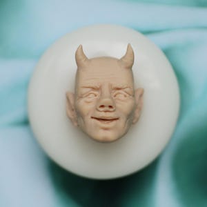 Puede incluir: Primer plano de una cara esculpida detallada de color marrón claro con cuernos. La cara sonríe y tiene una expresión ligeramente traviesa. La escultura está sobre un fondo blanco.