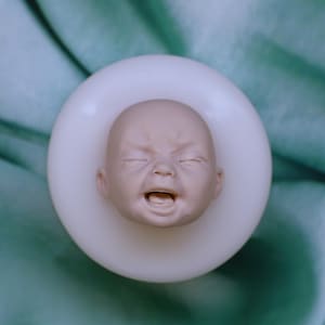 Puede incluir: Un molde de silicona para crear una cabeza de muñeca realista. El molde es blanco y presenta una impresión detallada de la cara de un bebé con los ojos cerrados y la boca abierta.
