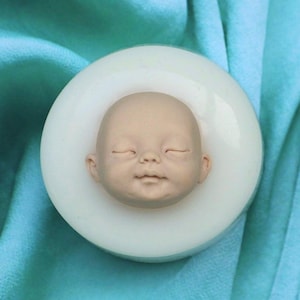 May include: A white silicone mould for making a baby doll head. The mould is round and has a detailed impression of a baby's face with closed eyes.