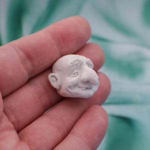 May include: A small, off-white sculpture of a smiling, elderly man's head. The sculpture has a large nose, detailed wrinkles, and a bald head. The figure is held in a person's hand against a soft, green background.