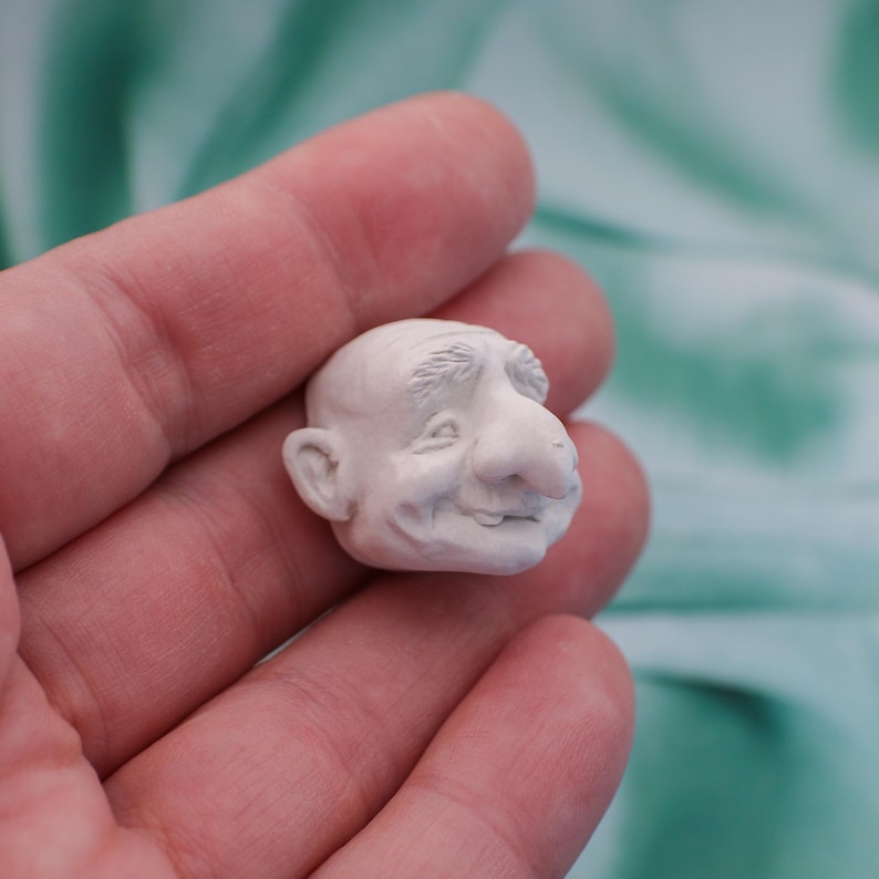 May include: A small, off-white sculpture of a smiling, elderly man's head. The sculpture has a large nose, detailed wrinkles, and a bald head. The figure is held in a person's hand against a soft, green background.