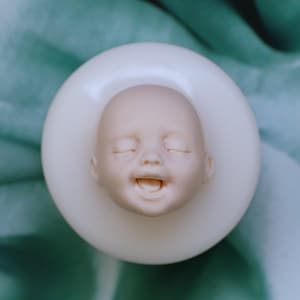 May include: A white porcelain mold of a baby's face with closed eyes and a smiling mouth. The mold is on a white circular base.