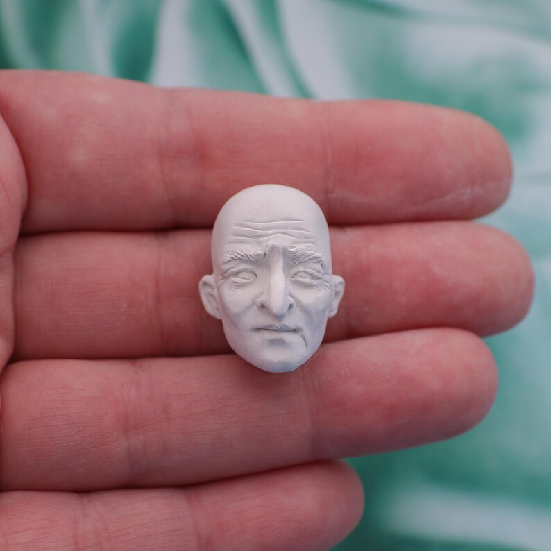 May include: A small, white, sculpted head of an elderly person with detailed facial features, including wrinkles and a bald head. The miniature sculpture is held in a person's hand, with a soft, blurred background.