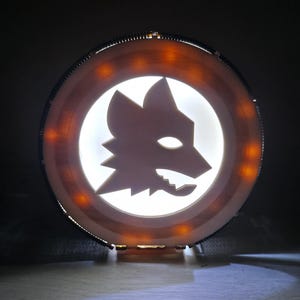 Può includere: Insegna luminosa circolare con l'emblema della testa di lupo bianco dell'AS Roma su sfondo bianco. La cornice in legno ha luci arancioni, creando un bagliore caldo. Oggetto decorativo.