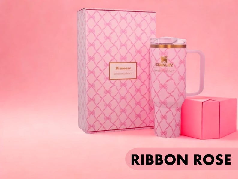 Puede incluir: Un vaso Stanley rosa con asa y tapa transparente, que presenta un patr&oacute;n repetitivo de lazos rosas. El vaso se muestra junto a una caja rectangular a juego con el mismo patr&oacute;n de lazos y una etiqueta de Stanley LoveShackFancy.
