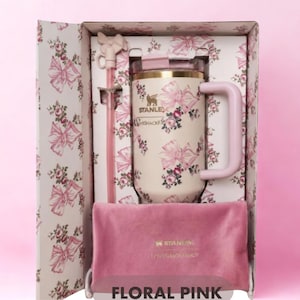 Puede incluir: Un vaso Stanley LoveShackFancy de color crema con un patr&oacute;n de flores y lazos rosas, que incluye una pajita rosa a juego con un adorno de lazo y una bolsa de terciopelo rosa, presentado en una caja decorativa con el texto FLORAL PINK abajo.