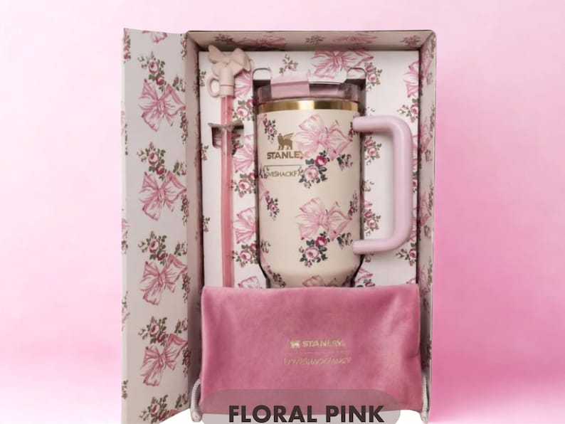 Puede incluir: Un vaso Stanley LoveShackFancy de color crema con un patr&oacute;n de flores y lazos rosas, que incluye una pajita rosa a juego con un adorno de lazo y una bolsa de terciopelo rosa, presentado en una caja decorativa con el texto FLORAL PINK abajo.