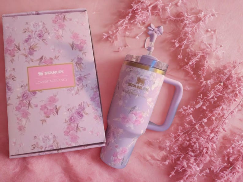 Puede incluir: Un vaso Stanley color lavanda con un patr&oacute;n floral y una caja a juego con el texto Stanley y LoveShackFancy. El vaso incluye un asa, una tapa transparente y un adorno para pajita con forma de lazo. El dise&ntilde;o floral presenta rosas de color rosa.