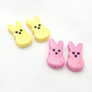 May include: Four small, bunny-shaped craft beads. Two are yellow with black dot eyes, and two are pink with glitter and black dot eyes. The beads have a hole for stringing.