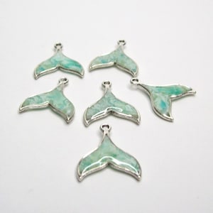 May include: Six silver-toned mermaid tail charms with a turquoise and white marbled design. Each charm has a small loop at the top for attaching to jewelry or other crafts. The charms are set against a white background.