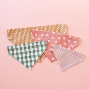 May include: A collection of five triangular bandanas in various patterns and colors. The bandanas include a green and white gingham, a pink floral, a tan leaf pattern, and a light purple polka dot design. The bandanas are arranged on a pink background.