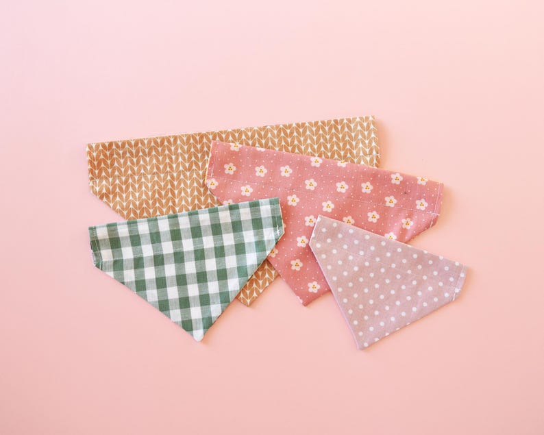 May include: A collection of five triangular bandanas in various patterns and colors. The bandanas include a green and white gingham, a pink floral, a tan leaf pattern, and a light purple polka dot design. The bandanas are arranged on a pink background.