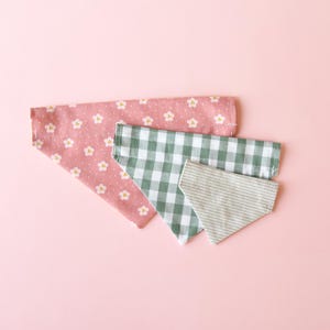 May include: Three dog bandanas in different patterns. The top bandana is pink with white floral designs. The middle bandana is green and white checkered. The bottom bandana is light gray with white stripes. All bandanas are triangular.