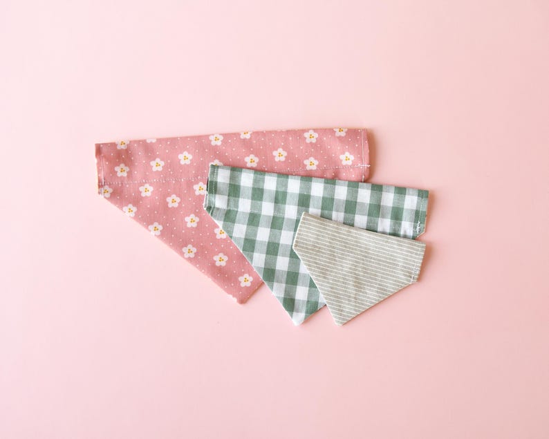 May include: Three dog bandanas in different patterns. The top bandana is pink with white floral designs. The middle bandana is green and white checkered. The bottom bandana is light gray with white stripes. All bandanas are triangular.