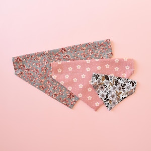May include: Four fabric bandanas in floral patterns, arranged on a pink background. One bandana is gray with red flowers, another is pink with white flowers, and a third is white with brown flowers. The fourth bandana is pink with white flowers.
