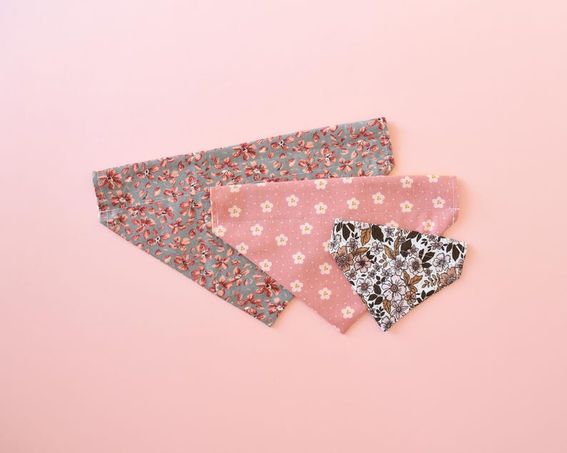 May include: Four fabric bandanas in floral patterns, arranged on a pink background. One bandana is gray with red flowers, another is pink with white flowers, and a third is white with brown flowers. The fourth bandana is pink with white flowers.