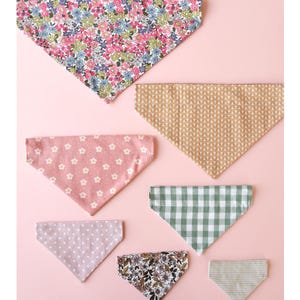 May include: A collection of seven triangular bandanas in various patterns and colors, including floral, polka dot, gingham, and striped designs. The bandanas are arranged on a light pink surface.