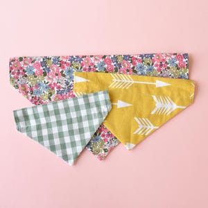 May include: Four colorful pet bandanas on a pink background. One is yellow with white arrows, another is green and white gingham, and two are floral with pink, blue, and white flowers.