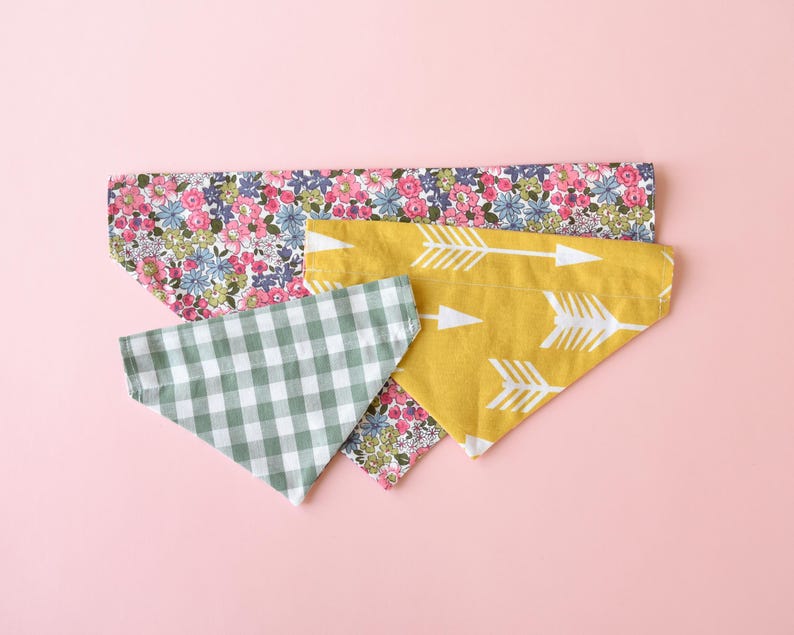 May include: Four colorful pet bandanas on a pink background. One is yellow with white arrows, another is green and white gingham, and two are floral with pink, blue, and white flowers.
