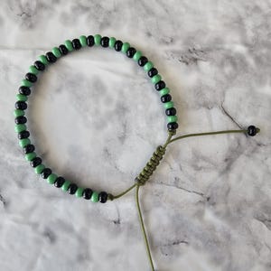 May include: A bracelet made of alternating black and light green beads. The adjustable bracelet has an olive green cord and a sliding knot closure. The beads are evenly spaced, creating a symmetrical design.
