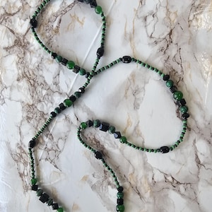 May include: A long necklace with a combination of green and black beads. The necklace incorporates round, oval, and geometric-shaped beads. The beads are strung on a green cord, creating a contrast against the black beads.
