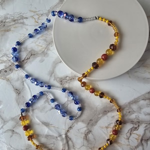 Peut inclure: Un collier de perles avec un dégradé de couleurs. Il présente des perles bleues, jaunes et marron de différentes formes et tailles, enfilées sur un cordon transparent. Le collier est présenté sur une surface circulaire blanche.