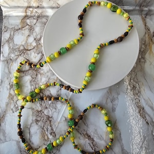 Op de afbeelding: Een lange kralenketting met een herhalend patroon van gele, groene en bruine kralen. De streng bevat een mix van ronde, gefacetteerde en cilindrische kralen in diverse tinten limoengroen, smaragdgroen, felgeel en donkerbruin hout.