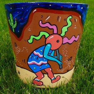 May include: A hand-painted terracotta flower pot with a whimsical design. A blue, pink, and green creature plays a flute, surrounded by stars and musical notes. The pot is decorated with a brown, yellow, and white background.