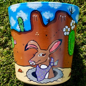 May include: Hand-painted terracotta flower pot with a desert scene. A brown rabbit with a purple bandana peeks out of a hole in the ground. The background features blue sky, white clouds, and green cacti.