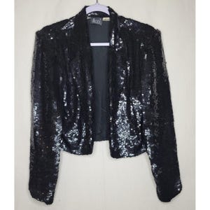 May include: Black sequin jacket with long sleeves and a notched collar. The jacket is fully covered in shiny black sequins. The label reads "MODI". The jacket is hanging on a purple hanger.