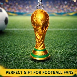 Puede incluir: Un llavero dorado con el trofeo de la Copa del Mundo, con un anillo y una cadena plateados. El llavero está sobre un campo verde con un balón de fútbol y un estadio de fondo. El texto en la parte superior dice "WORLD CUP TROPHY KEYCHAIN".
