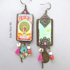 May include: A pair of rectangular drop earrings featuring bronze-toned metal hooks and filigree accents. One earring displays a colourful peacock graphic with the text PO SING, while the other shows a light green surface. Both include pink tassels and beads.