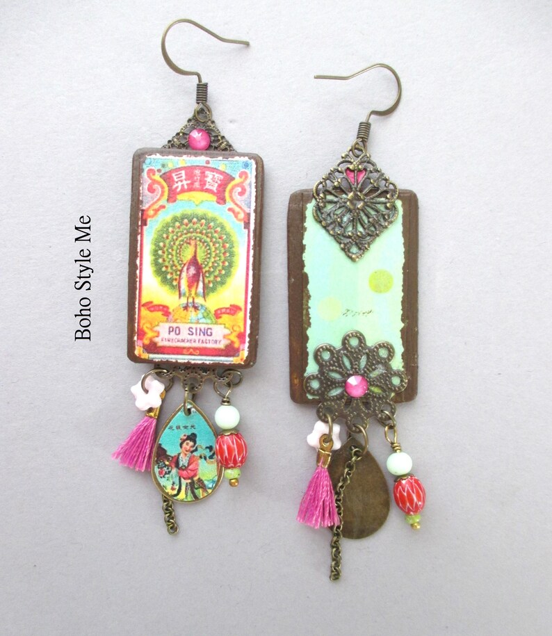 May include: A pair of rectangular drop earrings featuring bronze-toned metal hooks and filigree accents. One earring displays a colourful peacock graphic with the text PO SING, while the other shows a light green surface. Both include pink tassels and beads.