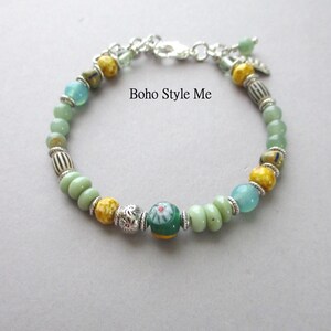 May include: A beaded bracelet featuring a mix of light green, yellow, and blue beads interspersed with silver-toned metal spacers and a central green bead with a floral pattern. The text Boho Style Me is visible. It includes a silver-toned clasp.
