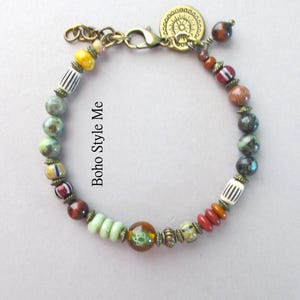 K&ouml;nnte beinhalten: Ein Perlenarmband mit einer Vielzahl von bunten Stein- und Glasperlen, darunter gestreifte, runde und scheibenf&ouml;rmige Elemente. Das Armband enth&auml;lt metallische Zwischenst&uuml;cke in Antikmessing-Optik, einen Karabinerverschluss und einen runden Anh&auml;nger. Der Text lautet Boho Style Me.