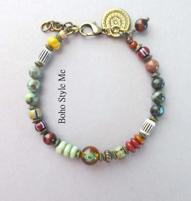 K&ouml;nnte beinhalten: Ein Perlenarmband mit einer Vielzahl von bunten Stein- und Glasperlen, darunter gestreifte, runde und scheibenf&ouml;rmige Elemente. Das Armband enth&auml;lt metallische Zwischenst&uuml;cke in Antikmessing-Optik, einen Karabinerverschluss und einen runden Anh&auml;nger. Der Text lautet Boho Style Me.
