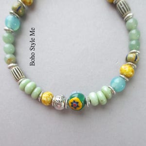 May include: A beaded bracelet featuring light green, yellow, and blue beads interspersed with silver-toned spacers and patterned glass beads. The text Boho Style Me is printed on the image. The design includes a central bead with a floral motif.