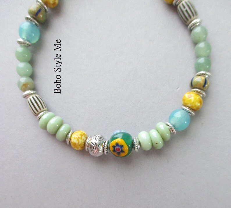 May include: A beaded bracelet featuring light green, yellow, and blue beads interspersed with silver-toned spacers and patterned glass beads. The text Boho Style Me is printed on the image. The design includes a central bead with a floral motif.