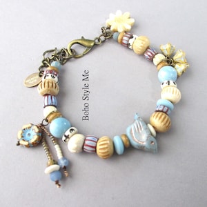 May include: A charm bracelet featuring a mix of light blue, cream, and striped beads, a bird-shaped charm, a flower-shaped charm, and a bronze-toned lobster clasp. A small oval tag with the text Boho Style Me is attached to the chain links.