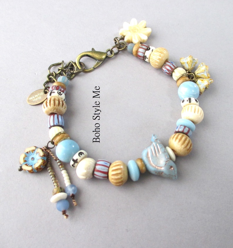 May include: A charm bracelet featuring a mix of light blue, cream, and striped beads, a bird-shaped charm, a flower-shaped charm, and a bronze-toned lobster clasp. A small oval tag with the text Boho Style Me is attached to the chain links.