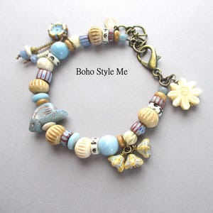 May include: A charm bracelet featuring a variety of beads in shades of light blue, cream, and tan, including a fish-shaped bead and flower-shaped charms. The bracelet has a bronze-toned lobster clasp and includes the text Boho Style Me.