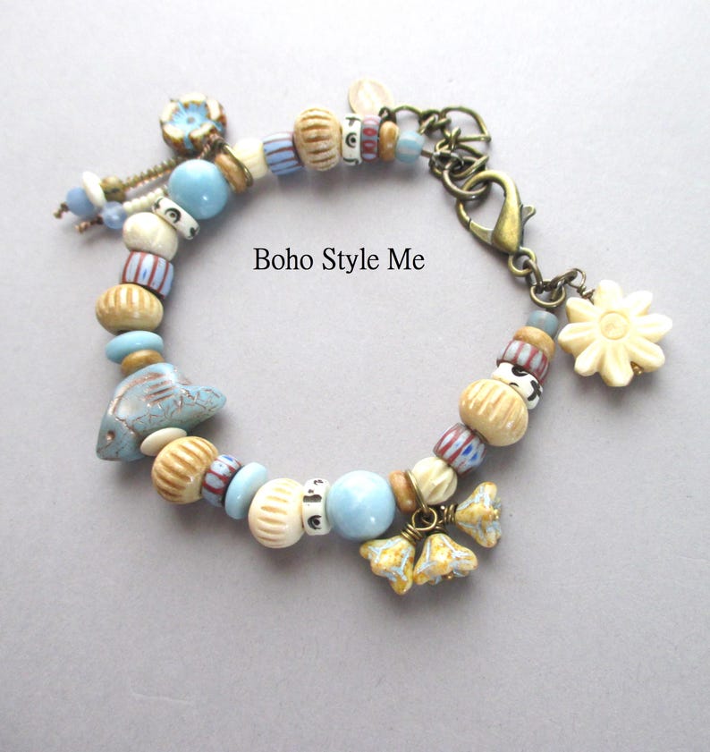 May include: A charm bracelet featuring a variety of beads in shades of light blue, cream, and tan, including a fish-shaped bead and flower-shaped charms. The bracelet has a bronze-toned lobster clasp and includes the text Boho Style Me.