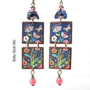 Puede incluir: Un par de pendientes colgantes con dos piezas cuadradas de madera apiladas con fondo azul oscuro y un estampado colorido de flores y mariposas. Los pendientes incluyen una cuenta azul oscuro en forma de campana arriba y una peque&ntilde;a cuenta rosa.