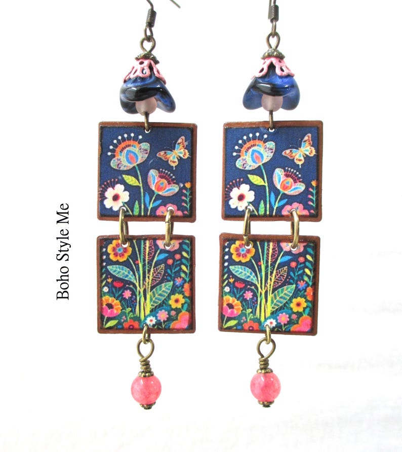 Puede incluir: Un par de pendientes colgantes con dos piezas cuadradas de madera apiladas con fondo azul oscuro y un estampado colorido de flores y mariposas. Los pendientes incluyen una cuenta azul oscuro en forma de campana arriba y una peque&ntilde;a cuenta rosa.