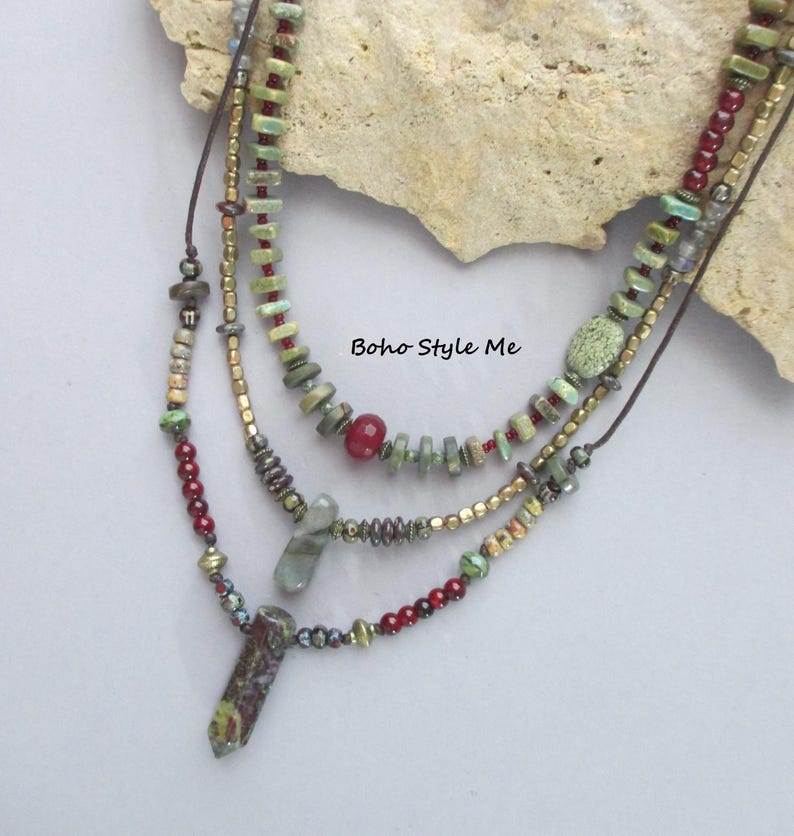Puede incluir: Un collar de varias hebras con cuentas verdes y rojas, detalles met&aacute;licos y dos colgantes de piedra. El collar incluye peque&ntilde;as cuentas cil&iacute;ndricas, cuentas rojas redondas y dos puntas de piedra alargadas. El texto Boho Style Me es visible.